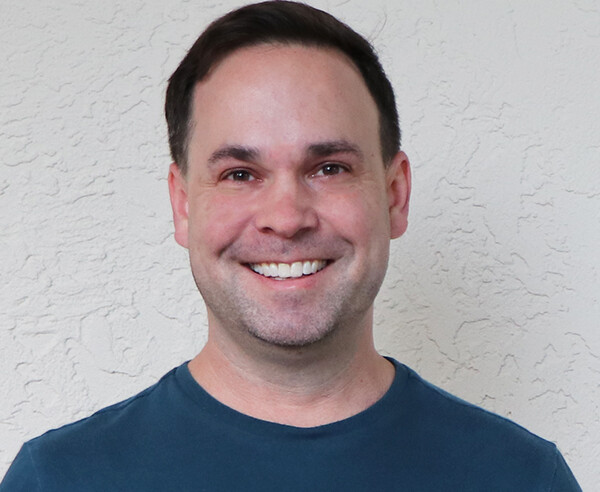 A man in a blue shirt smiling in front of a wall.