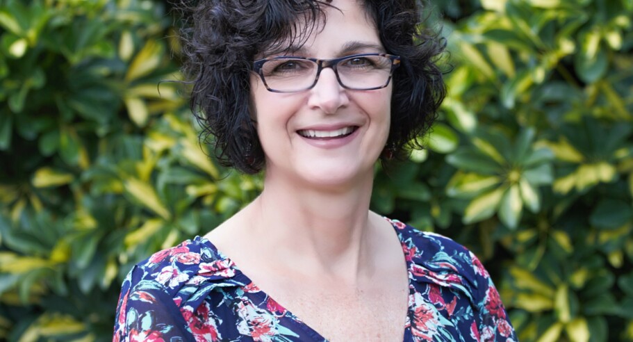 A woman in glasses smiling in front of bushes.