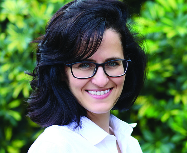 A woman in glasses smiling in front of bushes.