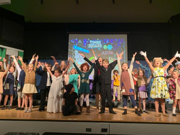 A group of children on stage with their hands up.