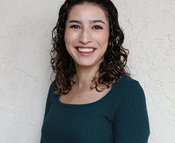 A woman in a green sweater smiles in front of a wall.