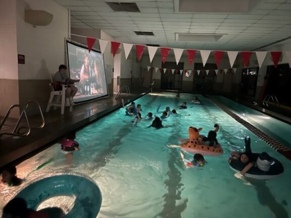 A group of youth floating in an indoor pool watching a movie.