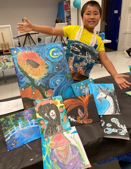 A young boy stands in front of a table full of paintings.