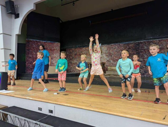 Children in colorful clothing are performing on a stage with a brick wall backdrop. Some are jumping, while others stand or kneel.