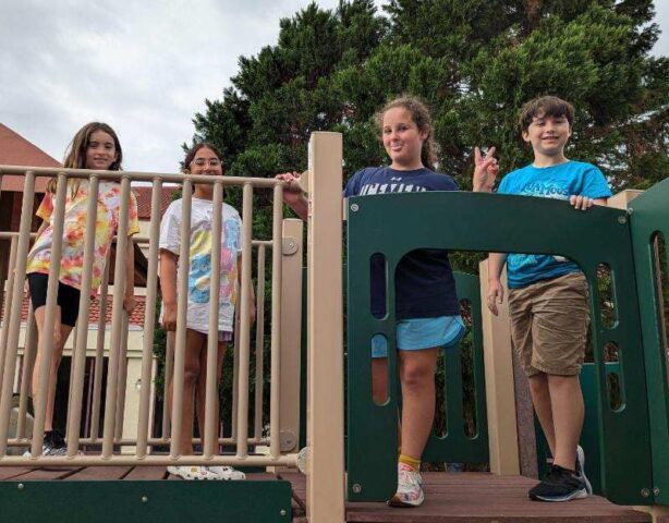 Four children stand on a playground structure, smiling at the camera. Trees and a building are in the background.