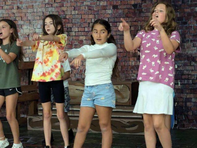 Five children stand in a row against a brick wall, performing a choreographed routine with their arms extended.