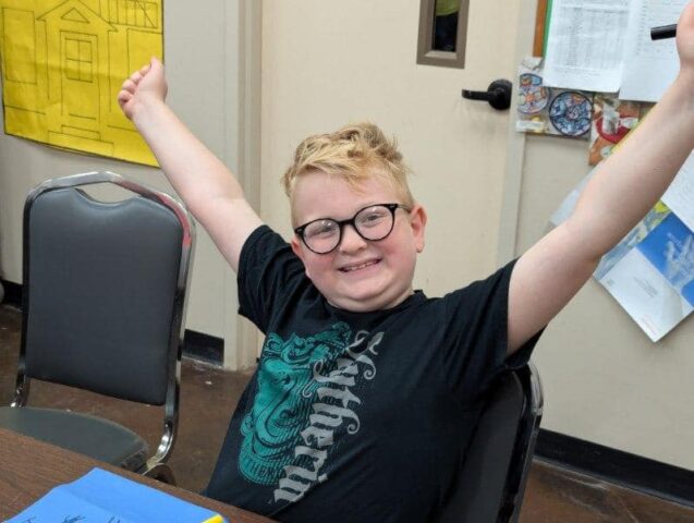 A child with glasses and a black t-shirt sits at a table, smiling and raising both arms triumphantly.