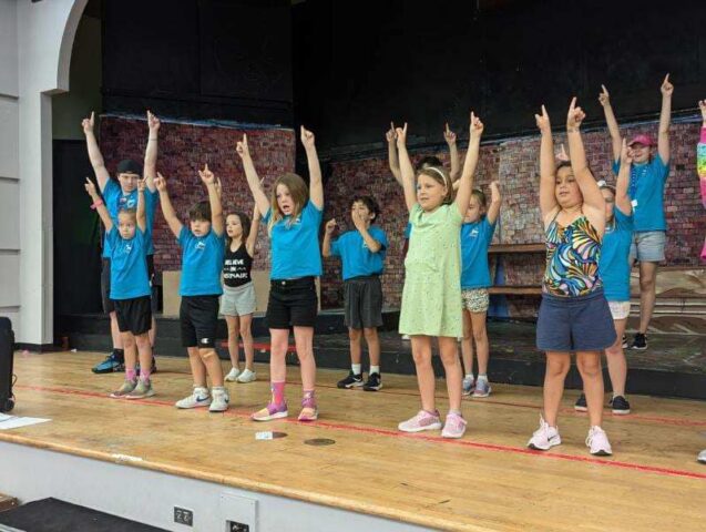 A group of children stands on a stage, raising their arms in unison, wearing casual clothes and appearing to rehearse a performance.