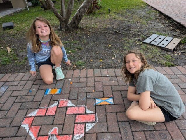 Two children are sitting on a brick path drawing colorful flags and designs with sidewalk chalk.