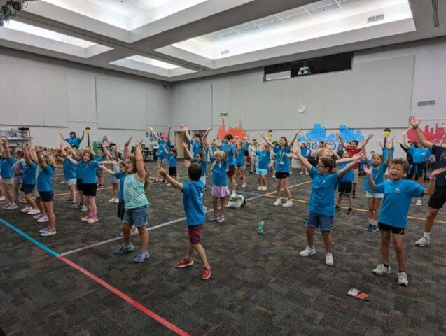 A group of children and adults are exercising in a large indoor room, all wearing blue shirts and raising their arms.