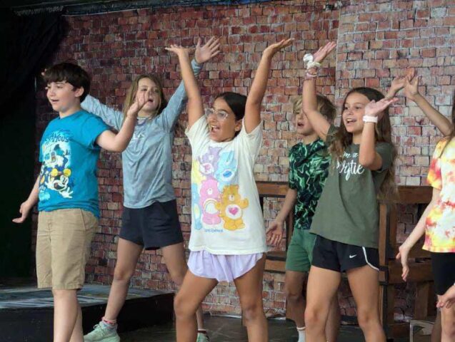 A group of children perform with arms raised on a stage in front of a brick wall.