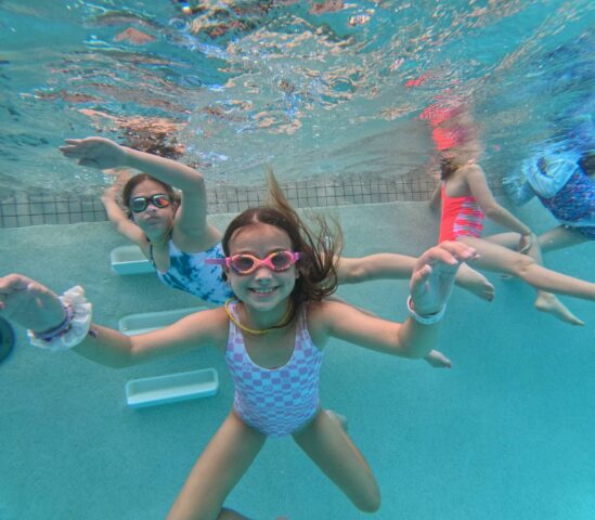 Children swimming underwater in a pool, wearing goggles and colorful swimsuits, with clear water and pool floor visible.