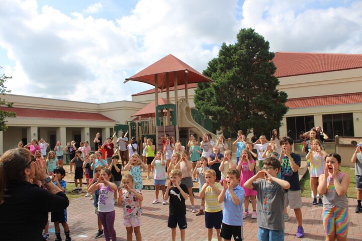 A large group of children and a teacher participate in an outdoor activity near a playground, in front of a building with a red roof, under a partly cloudy sky.