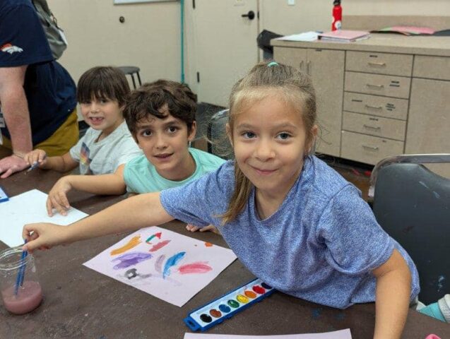 Three children are painting with watercolors at a table, smiling at the camera. Art supplies are scattered on the table.