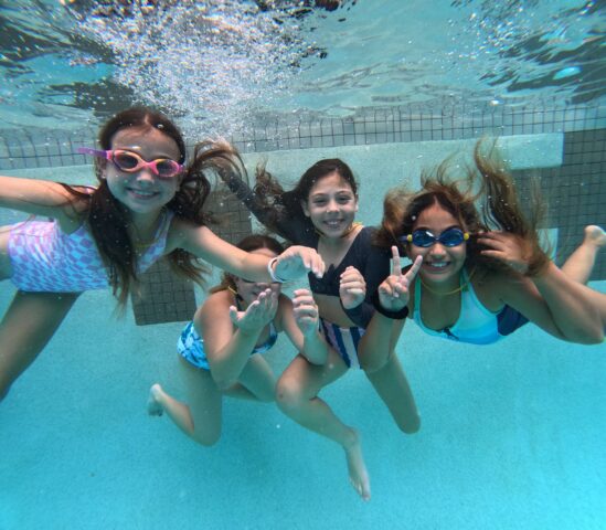 Four children swimming underwater, wearing goggles and swimsuits, smiling and making peace signs.