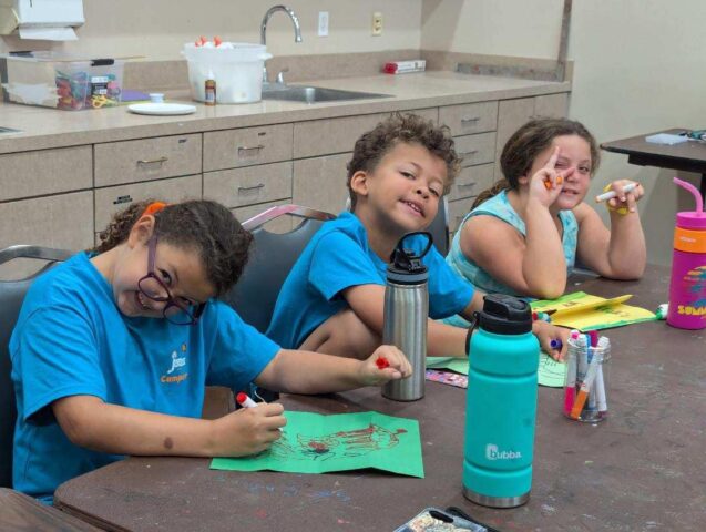 Three children sitting at a table coloring on paper, surrounded by art supplies and water bottles in a brightly lit room.