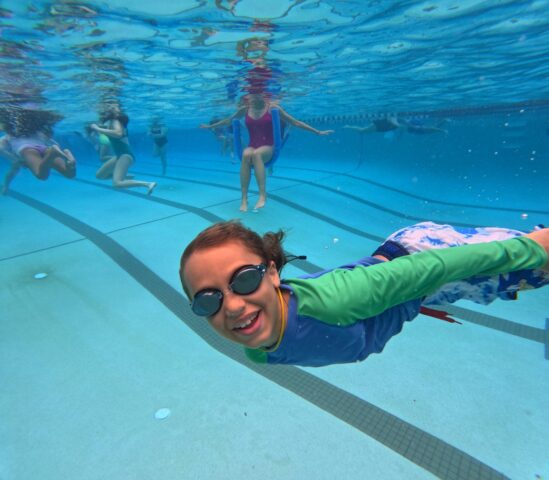 Child with goggles smiling underwater in a pool, surrounded by other swimmers in the background.