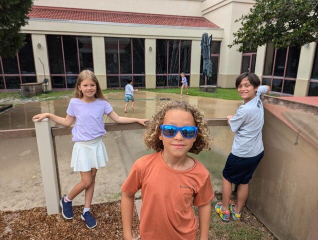 Three children stand by a railing near a wet pavement with a fountain in the background; one wears orange with blue sunglasses, another wears white and purple, and the third wears blue and crouches.