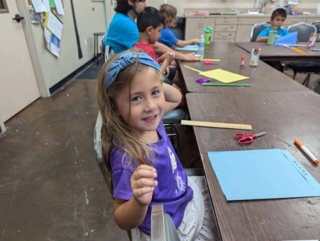 A young girl in a purple shirt sits at a table with paper and craft supplies, smiling at the camera. Other children are seated at the table, working on crafts.