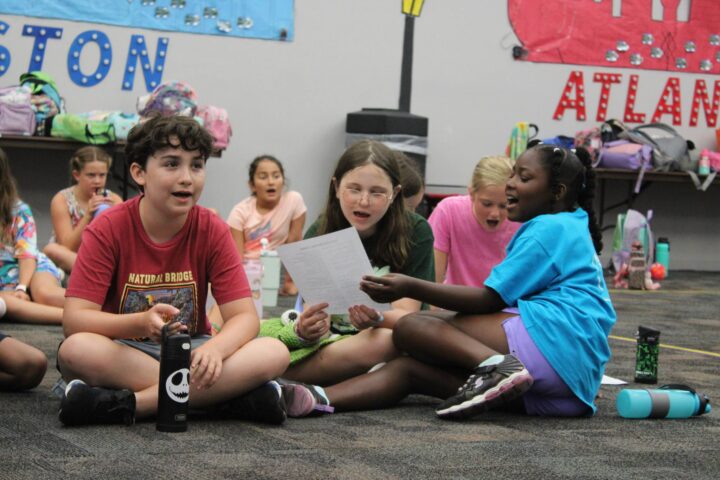 Children sitting on the floor in a classroom, reading from papers and surrounded by bags and posters on the wall.