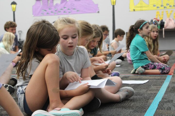 Children sitting on the floor in a classroom, looking at papers and listening. Walls are decorated with colorful posters.