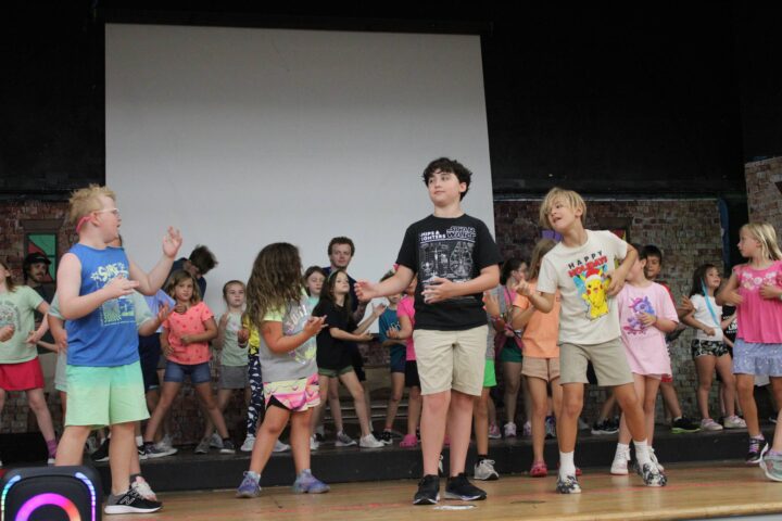 A group of children on stage, with some singing and others dancing. They are dressed casually and appear to be rehearsing a performance.