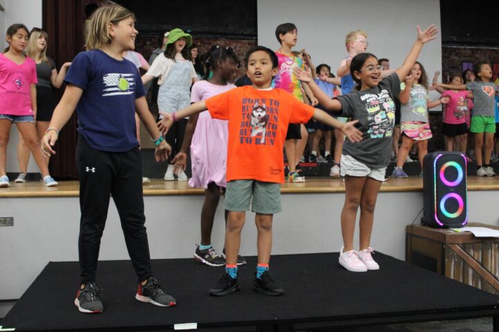 A group of children performing on stage, with three kids in the front striking a pose. They are surrounded by other children in colorful attire.