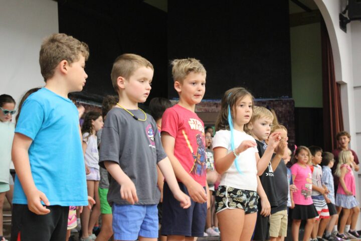 A group of children stands together on stage, appearing to participate in a performance. They are dressed casually and seem focused.