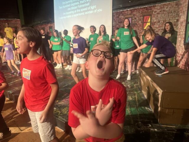 Children performing energetically on stage in colorful shirts, with one child in the foreground making a dramatic expression and pose. Text is projected on a screen in the background.