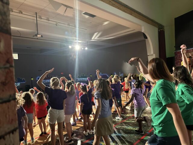 Children and adults wearing colorful shirts dance on a stage under bright lights in a large hall.