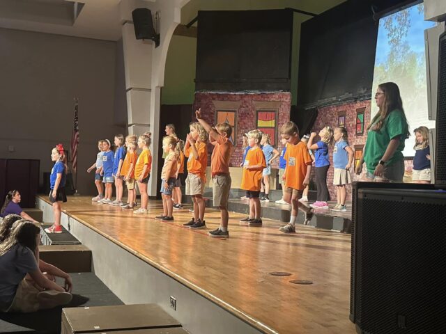 Children wearing orange and blue shirts perform on a stage with a backdrop of building facades. Adults stand nearby watching.