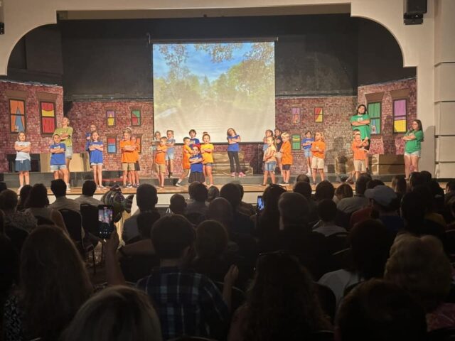 Children performing a play on stage with a projected backdrop and colorful costumes in front of an audience.