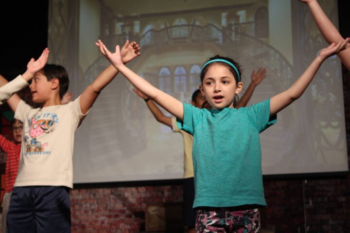 Children with arms raised are performing on stage with a projection of a grand staircase in the background.