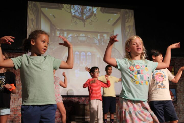 Children performing a choreographed dance on stage with a projected backdrop of a grand staircase.