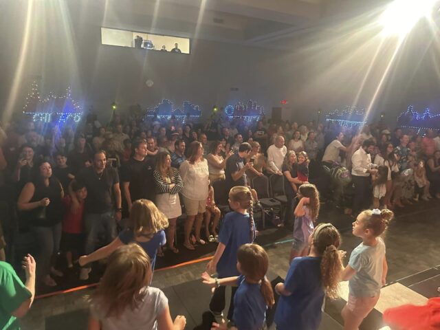 Children sing and dance on a stage in front of a large, applauding audience in a dimly lit room decorated with string lights.