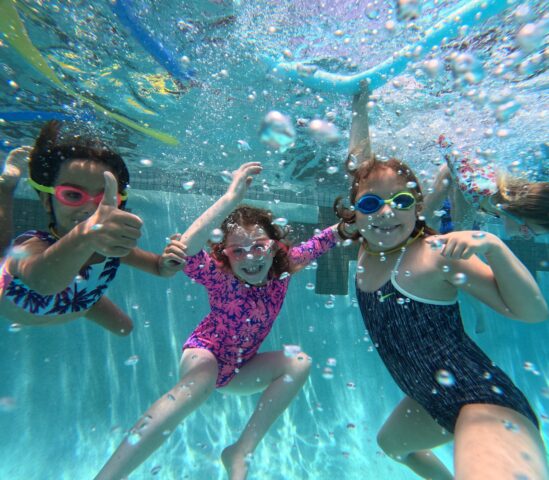 Three children wearing goggles and swimsuits swim underwater in a pool, smiling and giving a thumbs-up. Bubbles surround them, and colorful pool noodles appear in the background.