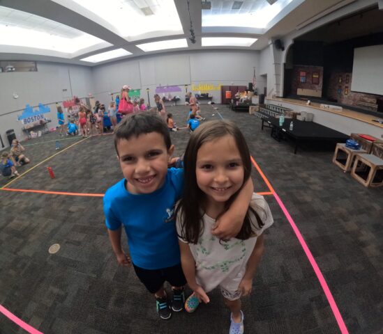 Two children smile at the camera in a gymnasium decorated with colorful murals. Other children and adults are visible in the background.