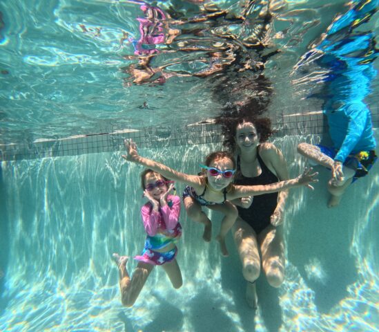 Three people underwater in a swimming pool, with two children wearing goggles and swimsuits, accompanied by an adult. Sunlight creates patterns on the pool floor.