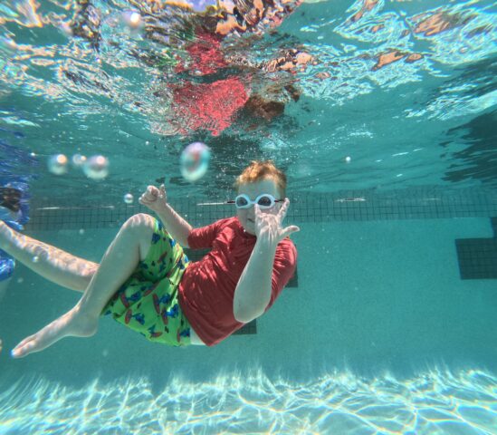 Child wearing goggles and colorful swimwear is underwater in a pool, surrounded by bubbles.