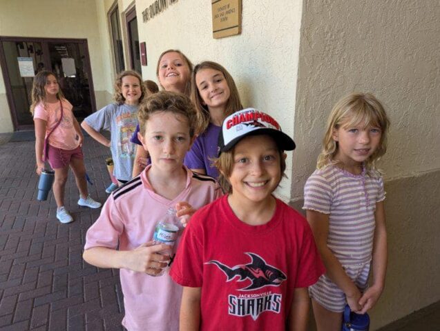 A group of children standing in a line outside a building, wearing casual clothes. One child in front is wearing a red shirt and cap. They appear to be smiling.