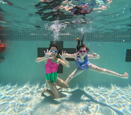 Two kids wearing goggles and bright swimsuits pose underwater in a pool, one holding up both hands, the other extending a leg.