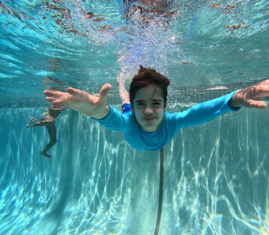 Child swimming underwater in a pool, reaching forward with both arms, while another person swims in the background.