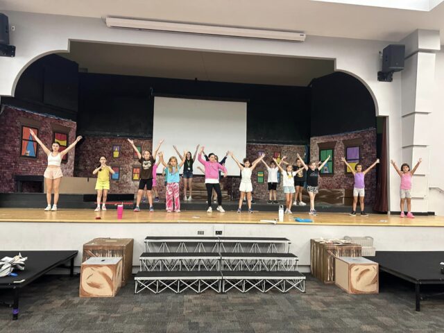 A group of children stand on a stage with arms raised, in front of a set designed to look like a brick building with colorful windows.