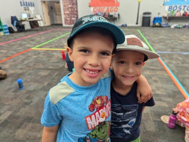 Two young boys smiling and standing together indoors, wearing caps and casual shirts. A colorful, spacious room is visible in the background.