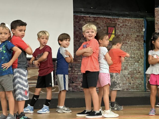 A group of young children standing in a line with arms crossed on a stage, some smiling.