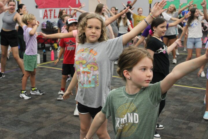 A group of kids in casual clothing practice dance moves in a large room with a carpeted floor.