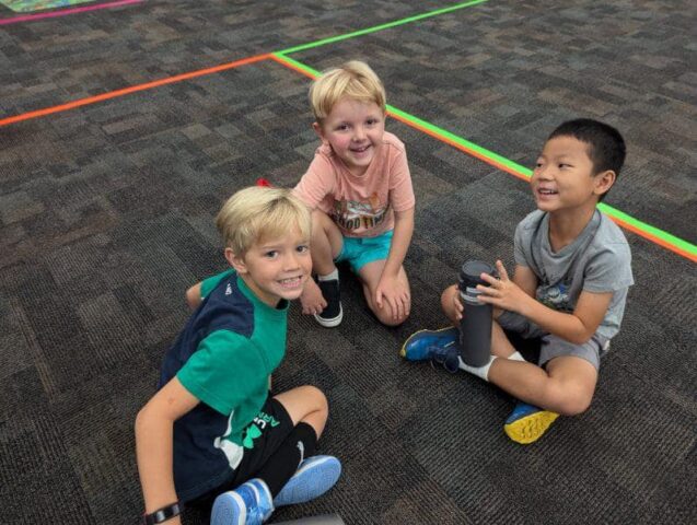 Three young boys sitting on a carpeted floor, smiling, with one holding a water bottle. They are surrounded by colorful taped lines.