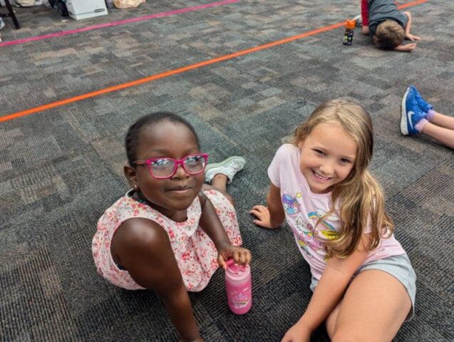 Two children sit on a carpeted floor. One has pink glasses and a floral dress, and the other wears a pink shirt with shorts. A child lies on the floor in the background.