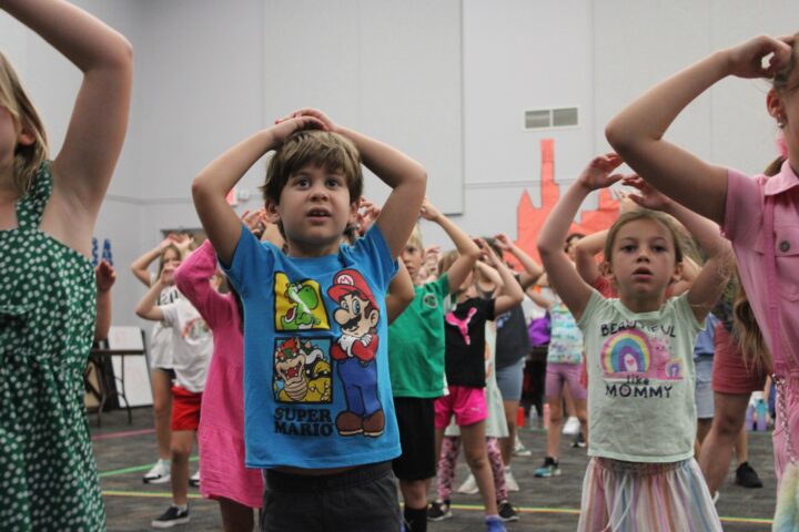 Children standing with hands on their heads in a group activity, wearing casual clothing, in a room with a patterned floor and white walls.