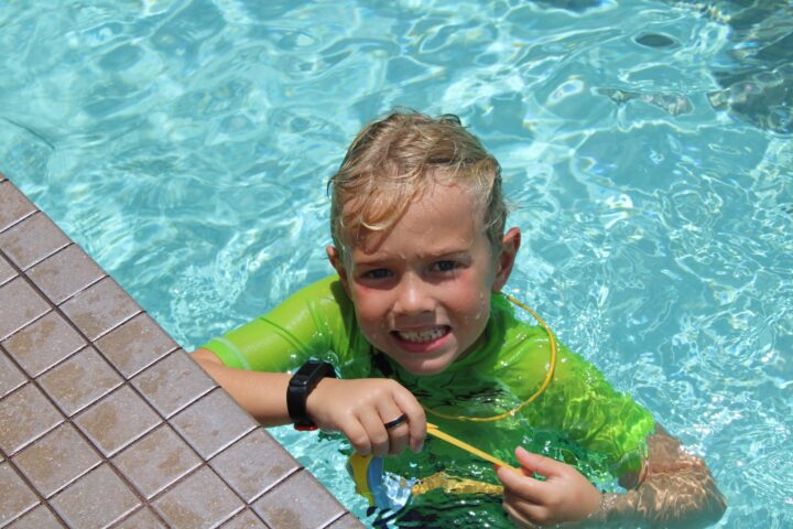 Child in a green rash guard smiles while standing in a swimming pool, holding onto the pool edge.